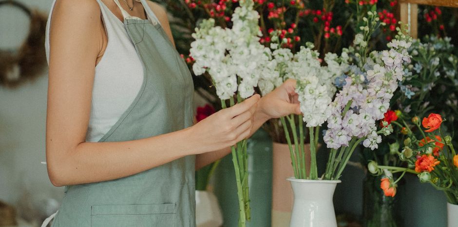 A person wearing a light grey apron over a sleeveless white top is arranging a bouquet of white, pale purple, and soft green flowers, which include stock and other floral varieties, in a tall white ce
