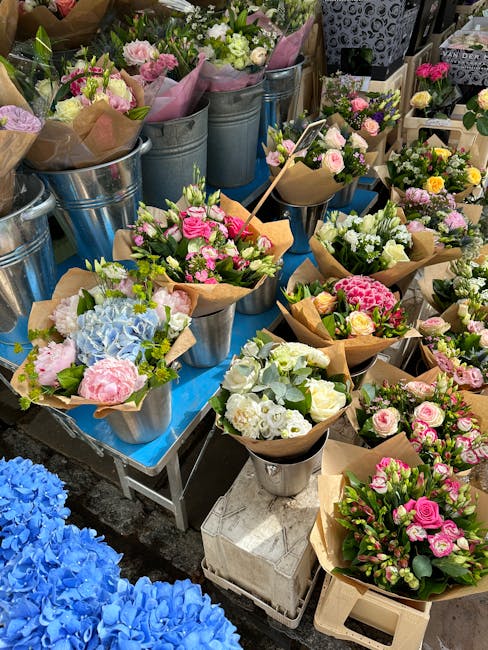 A vibrant display of fresh floral arrangements at a flower market, featuring pastel-colored roses, hydrangeas, and lilies wrapped in brown kraft paper and placed in small metal and glass vases. The fl