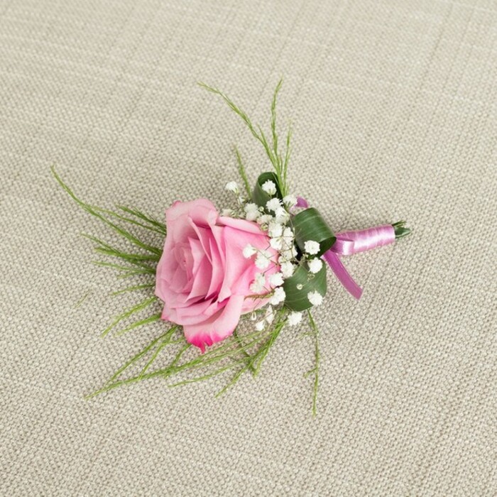 Resting on a softly textured, light beige linen background, a pink rose boutonniere is photographed from above in a way that reveals every careful detail, as if laid out on a dressing table in an Oxford townhouse just off Iffley Road before a ceremony begins. The central rose is in a beautifully open state, not yet fading, with velvety petals that spiral outwards from a tight blush-pink core. As the petals unfurl, the colour shifts into a slightly deeper pink near the outer edges, where the tips curl with a natural, gentle grace, suggesting freshness and quiet romance. Around the base of the rose, clusters of white baby's breath gather in soft, cloud-like tufts, their tiny, rounded blooms lending a light, airy texture and a traditional bridal feel that pairs effortlessly with the rose. From beneath the flower, slender blades of bright green, grass-like foliage fan out, fine and wispy, adding movement and a touch of wild garden charm, reminiscent of walks through University Parks in early summer. A single broader, darker green leaf is carefully folded and wrapped to cradle the stems, creating a structured, almost tailored collar that keeps the boutonniere compact and refined. The stems are tightly bound in a smooth satin ribbon in a soft pink tone, wrapped with precision and finished in a small, elegant bow whose short tails rest neatly against the linen. A slim, silver-toned pin is tucked at the base, ready to secure this floral accent onto a suit lapel for a groom, best man, or prom guest. The natural, even lighting casts delicate shadows around the rose and foliage, emphasizing the three-dimensional shape and the interplay between the plush petals, airy gypsophila, glossy ribbon, and woven fabric surface.
