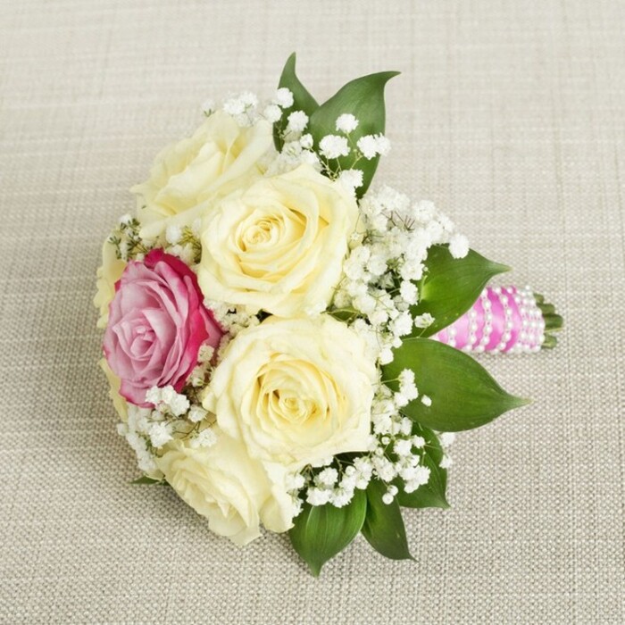 Displayed overhead on a softly textured, light beige fabric, this bouquet radiates understated elegance, graced by influences of Oxford's classic charm. At its heart are five to six creamy, pale yellow roses, petals cupped tightly in the bud and unfurling outward with youthful vigor, echoing the purity of new beginnings at university ceremonies and riverside weddings. Off-center to the left, one vivid magenta-pink rose instantly commands attention, injecting energy and individuality much like a brilliant poppy among the meadows of Port Meadow. Dainty white gypsophila, or baby's breath, is scattered liberally, infusing the bouquet with an ethereal, almost lace-like texture that contrasts exquisitely with the robust forms of the roses. Framing this ensemble are lush, glossy emerald leaves, their verdant surfaces catching the light to highlight the bouquet's depth and structure. The handle-meticulously wrapped in a bright pink satin ribbon-extends rightward, adorned elegantly with spiraled strands of shimmering pearls, giving the piece a sophisticated finish reminiscent of Oxford's elegant event spaces. Every element rests on the softly woven linen backdrop, ensuring the bouquet's subtle hues and striking contrasts take center stage. A beautiful reflection of romance and artistry, this bouquet captures the spirit of weddings, anniversaries, and special moments that echo through the historic streets and chapels of Oxford.