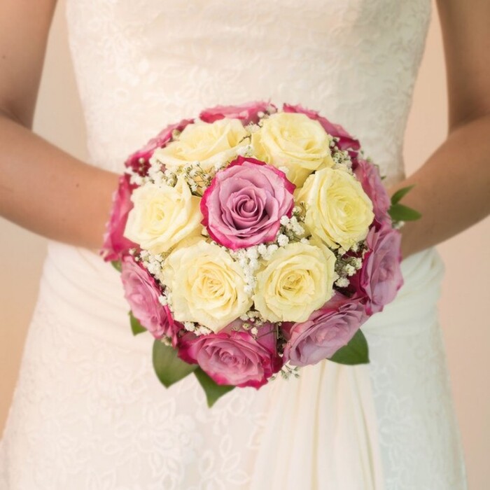 Against the softly blurred, sunlit backdrop of a classic Oxford setting, a bride's hands are seen holding a spectacular round bouquet at the centre of the frame-reflecting the elegance so often admired in the city's grand wedding venues. The bouquet brings together a medley of pale, creamy yellow roses mingling seamlessly with saturated fuchsia pink and dusky mauve roses, their petals impeccably layered for sumptuous texture and depth. Threaded throughout the arrangement are sprigs of delicate white baby's breath, adding a gentle, effervescent beauty reminiscent of mist along the banks of the Cherwell. Touches of dark green foliage emerge thoughtfully at the bouquet's edges, grounding the design and underscoring the freshness prized by Oxford's finest florists. The bride's forearms, bare and graceful, cradle the flowers at waist height, her flowing white gown providing a canvas of intricate lace detailing up top and smooth, flowing fabric below. Each element-petals, lace, and light-interacts harmoniously, as if curated for a celebration in the heart of Summertown or Jericho. Illuminated by even, natural lighting, the image conveys a mood of romance and joyful anticipation, serving as an inspiring reference for those seeking wedding floral arrangements with a distinct Oxford sensibility.
