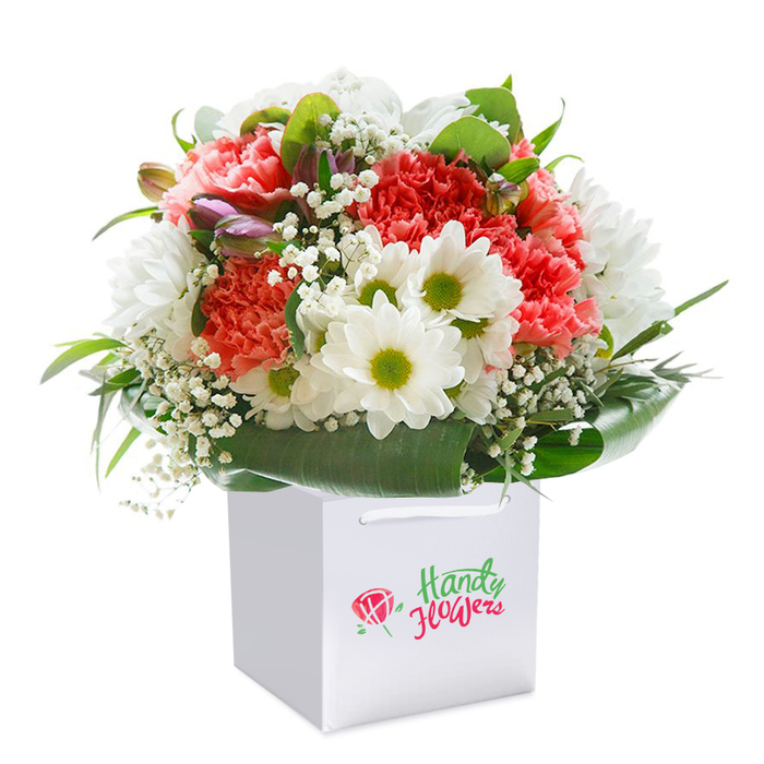 In this skillfully arranged floral bouquet, every petal and stem seems to celebrate the natural elegance of Oxford-each detail lovingly composed to showcase the artistry of local floristry. The bouquet's palette features soft coral and blush pink carnations, easily recognized by their layered, frilly petals, intermingled with energetic pops of deep red. Between these larger blooms, clusters of vivid white daisy chrysanthemums with zingy lime-green centers shine, casting a lively contrast reminiscent of radiant afternoons in South Parks. Airy sprays of baby's breath delicately punctuate the arrangement, adding gentle volume and softness to the overall design. Alongside, unopened purple-hued buds evoke a sense of anticipation-a quiet nod to the evolving beauty found throughout Oxford's Botanic Garden. Betwixt the blossoms, generous swathes of glossy, deep green foliage wrap the bouquet, their broad leaves forming a natural, supportive collar at the base. The ensemble stands upright inside a crisp white, cube-shaped gift bag, its sturdy ribbon handles peeking elegantly from the top. A sophisticated branding flourish graces the front-tasteful and unobtrusive, as befitting a high-end Oxford florist. The bouquet's rounded shape, vibrant colours, and tactile variety give it the air of a premium gift, suitable for occasions ranging from birthday gatherings in Jericho to a thoughtful gesture delivered to a College quad. Shot against a pure white background, this arrangement's freshness and cheerful energy radiate, making it a superb ambassador for Oxford's renowned flower delivery services and local floral artistry.