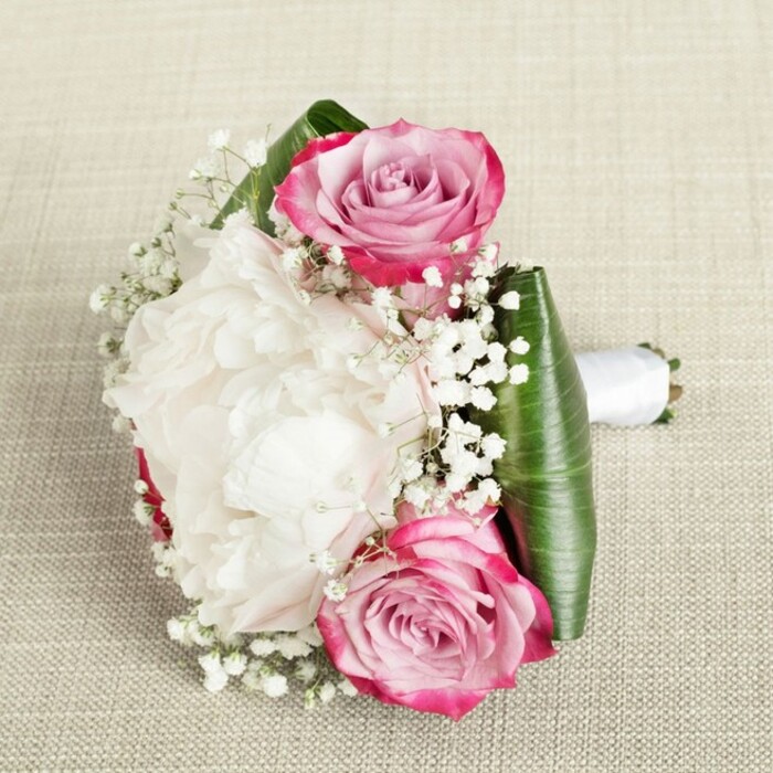 Seen from an artfully elevated angle, this sophisticated bridal bouquet pairs classical romance with Oxford's aesthetic grace. Against a neutral, tawny linen backdrop reminiscent of the city's centuries-old architecture, you'll find a lush centrepiece: a lavish white peony at full bloom, its ruffled petals inviting the eye with a tactile, creamy softness. Two radiant pink roses sit opposite each other-one near the top left, the other lower right-each reflecting a vibrant outer hue that mellows through gradients of blush and delicate lavender into their neatly whorled centres. The interplay between the peony's cloud-like fullness and the classic beauty of the roses is punctuated by graceful sprays of baby's breath, their tiny, alabaster blossoms lending a sense of lightness much like the gentle drift of blossom through Oxford's parks in early summer. Underpinning the bouquet is a collar of broad, dark green leaves, perhaps aspidistra or ti, precisely folded to cradle and elevate the floral artistry. The stems are bound in a pristine white satin ribbon, neatly extending to the right as a practical and stylish handle-ideal for the hand of an Oxford bride or bridesmaid. Soft, balanced light falls across each element, revealing the intricacies of petal, leaf, and ribbon alike, while the setting provides a warm, inviting neutrality. The bouquet radiates sophistication, serenity, and the promise of joyful moments-perfect for weddings, civil ceremonies, or refined occasions throughout Oxford.