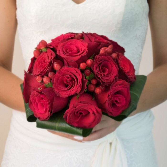 Illuminated by natural daylight reminiscent of an Oxford morning, this image reveals a bride-shown waist-up, centered outside one of Jericho's historic chapels-holding an elaborate classical bouquet. Her elegant white gown features a lace overlay with airy florals, a graceful V-neckline, and finely draped fabric at her side. The bouquet she cradles is impressive in its compact, rounded shape; at its core, a plenitude of rich crimson roses nestle tightly together, every spiral of petal subtly nuanced in shade and texture. Interspersed within are small, lively clusters of bright red hypericum berries, glinting in the light and adding a fresh, festive energy that recalls Oxford's spring markets. Broad, glossy green leaves, artfully folded into a structured collar, anchor the composition, balancing the passionate reds with a deep, natural calm. The vibrant hues of the bouquet contrast exquisitely with the brilliance of her gown. Every detail-from the tactile softness of the petals to the intricate play of light-reinforces the emotions of reverence, joy, and new beginnings. The gently blurred background-soft creams and ivories-keeps focus squarely on this showstopping arrangement, ideal for Oxford brides seeking both tradition and graceful modernity.