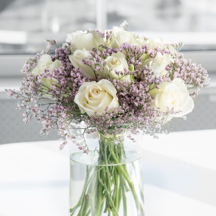 This detailed close-up shows an elegant sympathy bouquet that seems to glow softly in the light, as if positioned beside a quiet window in an Oxford terrace house. At the centre of the arrangement are numerous creamy white roses, their petals unfurling in perfect spirals that draw the eye gently inward. Each bloom is fully open yet still fresh, with velvety, slightly overlapping petals that create a sense of calm abundance rather than showiness. The roses are not a harsh white; instead they carry subtle ivory and off-white tones, lending warmth and tenderness, the sort of gentle beauty that feels appropriate for a condolence gesture. Surrounding and weaving between the roses are generous clusters of fine lavender and pale pink filler flowers, probably statice or limonium. These tiny, papery blossoms form a delicate halo of colour, creating movement and softness, like the muted pastels of an Oxfordshire sky at dusk beyond Port Meadow. Their light, misty appearance contrasts beautifully with the more substantial rose heads, adding both texture and depth to the design. All of the stems gather neatly into a tall, slender, clear glass vase filled with clean water, where the green stems are visible, straight and healthy, reinforcing a sense of freshness and care. The vase is placed on a pristine white surface that reflects the natural light, enhancing the bouquet's gentle luminosity. The background is out of focus, rendered in pale, neutral tones that keep attention firmly on the flowers. The overall mood is quiet, respectful, and soothing-a thoughtful floral tribute suitable for sending sympathy across Oxford, from Botley to Marston, when words alone are not enough.