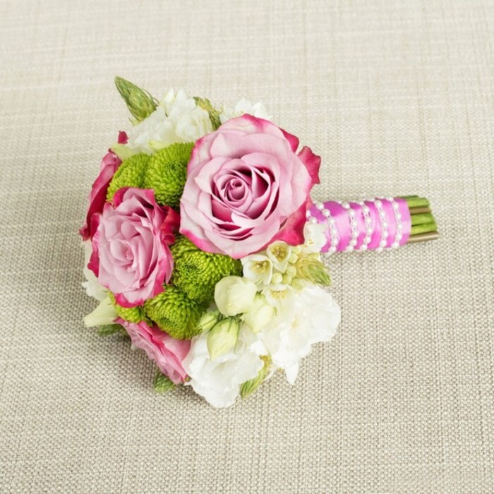 On a softly textured, light tan surface that looks like natural linen or burlap, a compact hand-tied bouquet is arranged horizontally, as though it has just been set down between photographs at an Oxford wedding. The neutral fabric, with its visible weave and gentle warmth of colour, forms a calm backdrop, allowing each stem and petal to be clearly defined. Dominating the centre of the design are three pink roses, two of them particularly prominent. Their petals are luxuriantly layered, loosely spiralling outward from tight centres, the inner petals a soft pastel pink that gradually deepens into richer, almost fuchsia tones at the edges. The very tips of the outer petals hold a suggestion of magenta, creating an elegant ombré effect that adds dimension and a sense of artisan care. Around the roses, bright lime green button chrysanthemums are grouped in tidy spheres, each little flower head composed of countless tiny, cushion-like petals. Their vivid apple-green hue injects energy and modernity, providing a crisp contrast to the romantic roses and evoking fresh spring growth in Oxford's University Parks. Threaded between the larger blooms are clusters of white and cream filler flowers - some with soft, cup-shaped petals reminiscent of lisianthus or freesia, others with small, starry blossoms and yellow centres that bring to mind waxflower or similar dainty varieties. These lighter elements lend airiness and gentle movement, as if the bouquet is breathing. Fine touches of darker green foliage peep between blossoms, grounding the colour palette and adding a botanical, garden-picked authenticity. The stems are gathered into a neat bundle, their cut ends aligned and slightly visible beyond a luxurious fuchsia pink satin ribbon wrap. This ribbon is smooth and lustrous, forming a polished handle suited perfectly to a bride or bridesmaid. Over the ribbon, a delicate string of white faux pearls winds diagonally in measured intervals, recalling vintage jewellery and enhancing the bouquet's bridal elegance. The lighting is soft and even, casting no harsh shadows, and the entire composition conveys intimacy and refinement - an ideal small bouquet for a bridesmaid walking through an Oxford college quadrangle, a civil ceremony at the Town Hall, or an anniversary dinner at a favourite restaurant near the Ashmolean. It feels thoughtfully crafted, romantic yet fresh, and ready to be carried or placed as a low, graceful table accent.