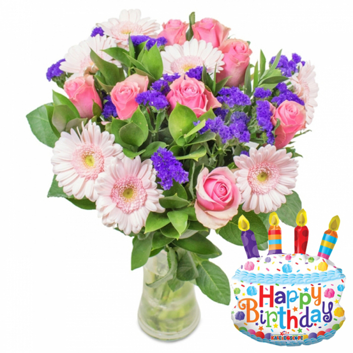 Set against a pristine white background, this festive floral composition reads like an invitation to celebration. The bouquet's dominant presence is a cluster of soft pink roses-each bloom carefully placed so that petals curl gracefully from a delicate blush toward slightly deeper pink rims-creating an overall impression of tenderness and warmth. Complementing the roses are pale pink Gerbera daisies whose broad, smooth petals radiate outward from soft yellow centers, adding a bright, cheerful geometry to the rounded rose forms. Sprays of vivid purple filler-likely statice or limonium-are woven throughout, their tiny, textured florets introducing a peppering of richness and subtle structural contrast. Small white blossoms are dotted through the grouping, lending moments of sparkle and additional textural variation. Surrounding foliage is abundant and glossy, a deep green backdrop that amplifies the flower colours and gives the bouquet a full, garden-picked silhouette. The stems sit in a clear glass vase with a slightly wider base where water and neatly trimmed stems are visible, reinforcing the sense of freshness and care that went into the arrangement. To the right-hand side, a metallic cake-shaped birthday balloon creates a playful counterpoint: frosted white surfaces, blue icing details, colorful sprinkles, and four multi-coloured candles with tiny yellow flame tips, all tied together by the multicoloured Happy Birthday script across the cake face; the small 'KALEIDOSCOPE' logo can be seen at the balloon's lower edge. This is the kind of bouquet a local florist in Oxford might prepare for a friend's birthday celebration near Christ Church Meadow or as a cheerful delivery to brighten a Summertown address.