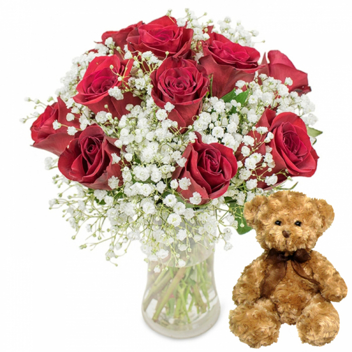 This carefully composed image features a classic romantic gift pairing, photographed against a smooth white background that keeps every focus on the flowers and the soft toy. On the left, a clear glass vase displays a generous bouquet of approximately one dozen deep red roses, each bloom full and luxurious. The petals are thick and velvety, layered in a spiral that draws the eye inward, with some roses completely unfurled and others only just reaching their peak, the way you might see them arranged on a counter in an Oxford florist near Cornmarket Street. Small touches of dark green foliage emerge between the blooms, but it is the interplay of colour that dominates: the intense crimson of the roses set against the crisp white of baby's breath. Clusters of Gypsophila weave lightly through the arrangement, their tiny, cloud-like flowers casting a softening veil over the rich reds, adding both visual lightness and a traditional, almost bridal charm. The vase is straightforward, cylindrical and crystal clear, revealing the straight cut stems inside and the clean water in which they stand, suggesting freshness and professional care. To the right of this romantic cluster, a medium-sized teddy bear sits upright, feet forward, like it has just been placed on a bedside table in a home off Cowley Road. Its fur is a gentle light brown, fluffy and slightly tousled, hinting at softness to the touch. The bear's black button eyes are rounded and bright, with a tiny embroidered nose and a delicately stitched smile giving it a comforting, affectionate expression. Around its neck, a slim dark brown satin ribbon is tied into a small bow, adding a finishing touch of understated elegance. The entire scene radiates warmth and sentiment, a visual echo of love notes, surprise deliveries across Oxford, and the quiet joy of being remembered on Valentine's Day, an anniversary, or a special birthday.