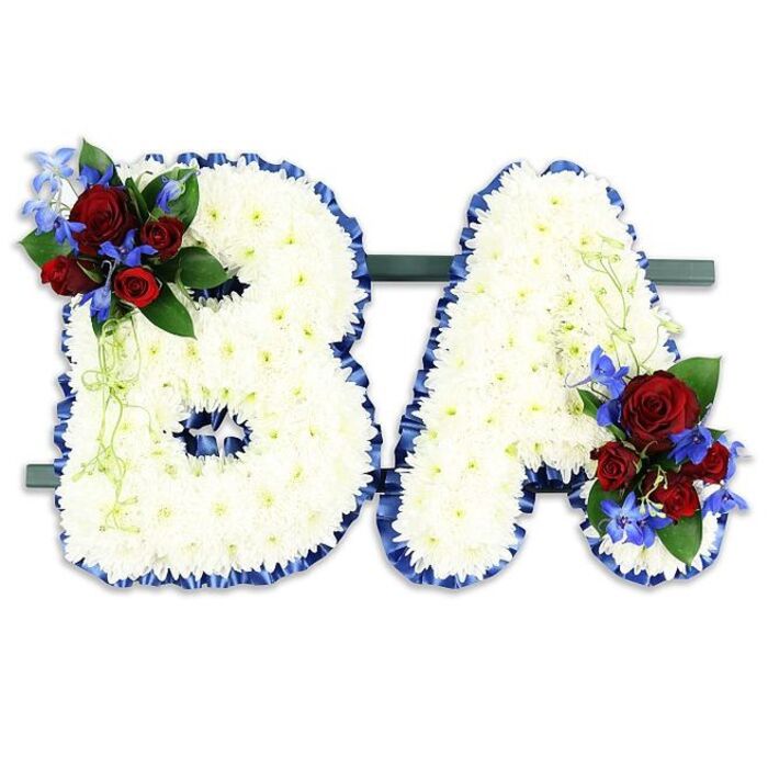 A dignified, thoughtfully composed floral memorial forms the initials BA, each letter densely covered with hundreds of immaculate white chrysanthemum blooms that create a soft, cloud-like texture and a sense of serenity. The contours of the B and the A are crisply defined by a ruffled band of royal blue satin ribbon, the rich colour providing a solemn frame against the crisp white studio background. On the upper left curve of the B a small, concentrated spray of deep red roses brings warmth and sentiment; varying stages of bloom add life and depth, their subtle fragrance tempered by the cool, intense blue spikes of delphinium-style flowers woven through the cluster. A sympathetic mirror cluster adorns the lower right of the A, balancing the composition and reinforcing the visual narrative of remembrance. Dark green foliage provides a supportive backdrop to each floral accent while pale green trailing tendrils soften the arrangement's edges and introduce an elegiac, almost whispering motion. The letters are secured side-by-side on a discreet dark-green rectangular base, a shallow stand designed for stable presentation at a chapel or service. Under gentle, diffused lighting the white chrysanthemums glow quietly, the satin ribbon catching highlights, and the red and blue accents read as focal points of affection and courage. This bespoke sympathy piece speaks to both personal connection and ceremonial grace, suitable for memorial services in and around University Parks, St Giles', or Port Meadow where families seek a carefully personalised tribute.