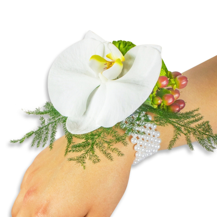 A beautifully crafted wrist corsage is displayed on a light-toned wrist against a pristine white background, designed to foreground form, colour and tactile contrast. The focal element is a large white Phalaenopsis orchid, its broad, velvety petals arranged like a quiet fan; the orchid's throat reveals a luminous yellow centre with touches of deeper orange, creating a warm focal point among the pale petals. Beneath the bloom, sprays of delicate asparagus fern unfold in lively green, their feathery texture creating a soft, organic frame that gives the composition depth. To the upper right of the bloom, a tight cluster of Hypericum berries provides a textural accent and a gentle colour gradient from light green through blush to a deep cranberry-red, while a small patch of ruffled lime-green foliage adds brightness and sculptural contrast. The arrangement is fastened on a classic multi-strand bracelet of tiny white pearls that wraps the wrist with understated glamour, the pearls' cool luster balancing the botanical tones. Set in soft, diffuse light - the kind of light that filters through Botanic Garden leaves or along Oxford's High Street on a calm afternoon - the corsage reads as both timeless and contemporary. It speaks to ceremonial moments: weddings, proms, anniversaries or graduation processions past the Covered Market. The image emphasises craftsmanship, freshness and wearability, highlighting the interplay of textures from smooth orchid petal to delicate fern frond and glossy berry, finished with a refined, wearable elegance suited to special occasions.
