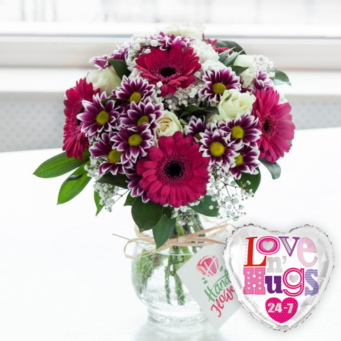 A richly detailed bouquet brimming with purple, magenta, and soft white blooms takes centre stage, as if waiting on a hallway console table in an Oxford Victorian terrace, ready to welcome someone home. The flowers are gathered in a classic, rounded clear glass vase, the transparent sides showing off an orderly cluster of green stems nestled in fresh water. Standing tallest and most dramatic are the large gerbera daisies in a vibrant fuchsia-magenta shade; their silky petals radiate out in perfect circles around dark, inky centres that give each flower a dramatic, almost graphic presence. Interwoven among them are generous sprays of two-tone chrysanthemums, each bloom a miniature starburst of deep purple fading to clean white at the tips, with bright yellow centres that glow warmly. Creamy white roses, some still in bud and others slightly opened, are tucked between the brighter flowers, their layered petals adding a classic, romantic softness to the arrangement. Fine clusters of white gypsophila float in between, like tiny clouds or lace, gently blurring the boundaries between bloom and foliage. The outer edges of the bouquet are framed with fresh, glossy greenery, lending structure and a natural, garden-fresh feel that would look at home after a stroll through University Parks. The vase itself is cinched at the neck with a length of rustic, natural twine tied in an uncomplicated bow. From it hangs a small, rectangular white tag, beautifully simple, showing a stylised pink rose icon and green handwritten-style text that suggests the flowers have been thoughtfully composed. Just to the right of the vase rests a heart-shaped foil balloon edged in shining silver. Its white centre is covered with playful, patterned letters spelling Love n' Hugs 24-7 in bright pinks, purples, and blues, echoing the cheerful colours of the bouquet. The softly blurred, bright background hints at daylight filtering through a nearby window, creating an uplifting atmosphere that suits anniversaries, birthdays, or same-day love-filled deliveries across Oxford, from Botley to Marston.