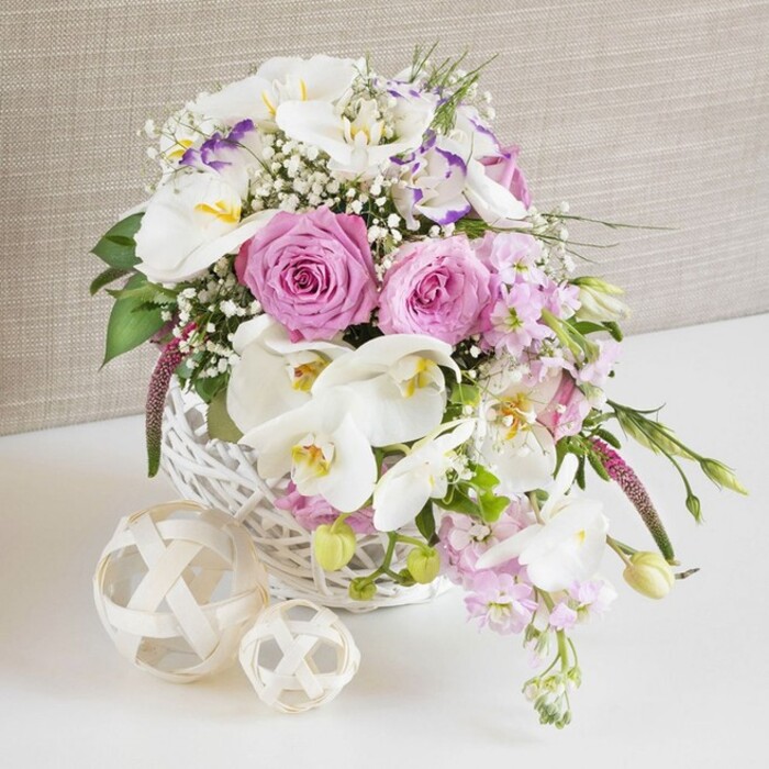A beautifully composed floral centrepiece overflows from a round white wicker basket, styled with the ease and refinement of a wedding reception table in the heart of Oxford. The arrangement glows with pastel tones: fully unfurled pink roses open wide to reveal their classic spiral centres, each petal delicately cupped and slightly curled at the edges, giving the piece a timeless, romantic character. Intertwined among the roses are striking white Phalaenopsis orchids, their broad, satin-smooth petals almost luminous against the softer blooms, with golden-yellow and soft peach centres that bring a hint of exotic elegance. Around them, ruffled white Lisianthus, some petals brushed with a fine line of lavender-purple, introduce subtle depth and movement, while stems of pale pink stock flowers emerge from the basket and gently cascade downward, elongating the design with a graceful flow. Fine threads of white baby's breath create a lace-like veil across the flowers, softening transitions and adding a whisper-light, airy quality. Glossy green foliage and newly forming buds slip between the blossoms, providing structure and a fresh garden feel, as though gathered from a well-tended border near the Oxford Botanic Garden. The basket rests on a crisp white surface, next to two decorative orbs fashioned from pale, woven natural fibres that echo the texture of the wicker and add an understated, organic touch. Behind, a neutral, light beige fabric backdrop, reminiscent of linen, blurs softly, ensuring that all attention stays on the flowers. The image evokes warmth, celebration, and thoughtful craftsmanship, making it easy to imagine this centerpiece enhancing a bridal table in Oxford's historic colleges, a special family dinner in Summertown, or an intimate anniversary brunch at home.