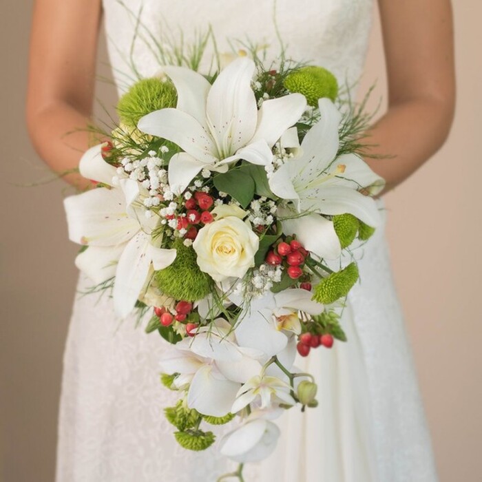 Highlighted in a close, intimate photograph, a bride in Oxford is depicted holding an exquisite cascading bridal bouquet, perfectly arranged to capture both classic beauty and fresh, contemporary charm. The dominant feature is several large, milk-white lilies, whose open trumpets are delicately patterned inside with fine russet freckles, forming an elegant focal point. Tucked at the heart is a perfectly coiled, creamy rose symbolising timeless sophistication-a signature touch for weddings throughout the city. The bouquet gains playful texture from spherical lime-green chrysanthemums, offering a nod to the dynamic energy of Oxford's bustling High Street. Bright red clusters of hypericum berries nestle amid the blooms, bringing a warm and festive contrast reminiscent of autumn strolls through South Park. Gentle baby's breath blossoms weave between the larger flowers, their tiny white petals lending a sense of airiness, while the bouquet's dramatic cascading shape is achieved with the downward flow of pristine white Phalaenopsis orchids, echoing the gentle tumble of water over Magdalen Bridge. Framing the arrangement, finely arched foliage and slender blades of grass evoke the wild, untamed feel of nature reserves like Shotover Country Park. Set against the soft, blurred fabric of the bride's lace gown, every component is rendered in rich, tactile detail. A true testament to expert floristry, this bouquet would make a luxurious statement at any Oxford wedding, reflecting the city's unique blend of tradition, artistry, and joyful celebration.