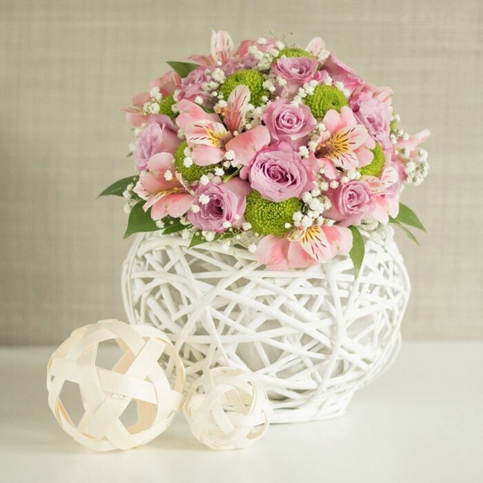 Commanding attention as a table arrangement worthy of Oxford's finest gatherings, this elegant, rounded bouquet is a masterful harmony of soft pastels and lively greens. Exquisite pastel pink and lavender roses, in the prime of bloom, are lovingly set at the heart of the arrangement, their plush petals forming gentle whorls. Graceful stems of pink alstroemeria-distinguished by slender petals and delicate internal markings-cascade naturally, woven amongst clusters of vibrant green button chrysanthemums whose spherical shapes offer a cheerful, modern contrast. White sprigs of dainty baby's breath are deftly layered throughout, imbuing the whole tableau with a light, floating airiness reminiscent of spring mornings along the Thames in Oxford. Deep green leaves emerge at intervals, introducing natural depth and structure to the composition. The flowers are cradled within a striking white vase, its intricate open-weave design reminiscent of handwoven baskets often seen in Covered Market stalls-substantial, rounded, and uniquely contemporary. Flanking the main display, two decorative spheres in pale, woven rattan add organic texture and visual interest, one slightly larger and more prominent, the other smaller and set forward, echoing the overall harmony of the design. The neutral, softly textured beige background ensures every bloom and detail shines, while the light-drenched surface suggests freshness and cleanliness. This floral centerpiece is an inspired choice for wedding receptions, anniversary tables, or celebratory events across Oxford, seamlessly blending classic English garden flowers with innovative, handcrafted accents and the hallmark finesse of a trusted local florist.