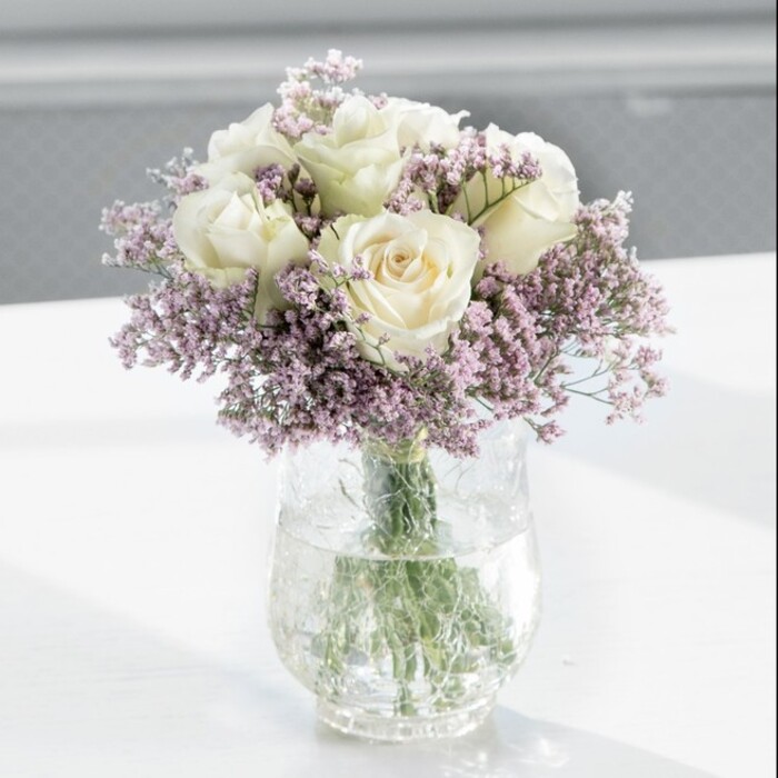 In this image, a lush, dome-shaped bouquet of creamy white roses and pale lavender statice stands tall in a clear cylindrical vase, evoking the gentle poise you might find in an elegant townhouse off Banbury Road in Oxford. The roses, roughly a dozen in number, are the undisputed focal point: each one perfectly fresh, petals smooth and velvety, curving outward from a softly cupped centre. Their tone is not a harsh white but a creamy, almost ivory shade that feels warm and welcoming, catching the light in such a way that each petal edge seems delicately highlighted. Threaded between the roses, sprays of fine-stemmed statice spread out like clouds of miniature blossoms. The flowers are a muted lavender-purple, their slightly papery, clustered blooms forming a light halo around the round, plush heads of the roses, adding dimension and a discreet pop of colour. Together they create an impression of gentle movement, as if a breeze had just passed through a garden border. The stems are cut evenly and held neatly within the vase, visible through the crystal-clear water that fills the lower half of the glass and throws back faint reflections on the white surface beneath. Soft daylight, similar to a calm mid-morning glow drifting through an Oxford studio window, pours in from one side, illuminating the blooms and leaving the background softly blurred, almost abstract. The arrangement feels perfectly at home as a centrepiece for a small wedding breakfast, a refined sympathy gesture, or a housewarming gift delivered across Oxford by Flowers Oxford, embodying that subtle mix of modern restraint and timeless romance that many local clients appreciate. It suggests a light rose fragrance and a soothing atmosphere, ideal for moments that call for both beauty and quiet reflection.