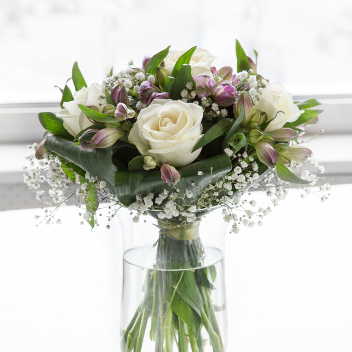 The photograph shows a tranquil, carefully crafted sympathy bouquet displayed in a cylindrical clear glass vase, standing in a pool of soft, natural light that could easily be streaming through an Oxford terrace window near Iffley Road. The arrangement is lush yet restrained, with creamy white roses forming the main focal point. Each rose is in that ideal stage between bud and full bloom, the petals rolled back in smooth, velvety layers that suggest gentleness and calm. Weaving around and between the roses are stems of alstroemeria in tones of lavender, dusky mauve, and hints of fresh green, their trumpet-shaped flowers offering a quiet contrast in colour while maintaining an overall soothing palette. Fine sprays of white baby's breath dance throughout the bouquet, occupying the small spaces and creating a light, airy veil that softens the transitions between blooms and foliage. The greenery is abundant and rich in tone, with glossy, deep green leaves and slender, upright stems forming a supportive framework that holds everything in balance. Through the clear sides of the vase, you can see the green stems immersed in clean water, straight and neatly cut, adding to the sense of freshness and care in the floristry. The background is almost entirely white and gently diffused, with no visual distractions, emphasising the bouquet's shape and harmonious colours. The overall impression is one of quiet elegance and sincere compassion, the sort of arrangement you might send across Oxford-from Botley to Marston-as a thoughtful gesture of sympathy, to sit beside a framed photograph or on a simple wooden table during a time of reflection.