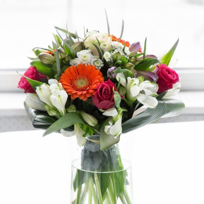 A scene of refined harmony unfolds in this expertly designed Oxford bouquet, where colour, structure, and emotion exist in perfect balance. Creamy gerberas create a gentle halo against which brilliant orange centers gleam and flicker, recalling festive lanterns strung through the city on celebratory nights. The magenta roses interspersed among them lend a luxurious depth, each tightly layered petal hinting at stories untold. Sprigs of ivory alstroemeria and fresh green leaves layer the arrangement, their textures echoing the lush, garden-fresh feel of Oxford's botanical treasures. Generous, paddle-shaped aspidistra leaves curve gracefully around the stems, forming a verdant collar much like the sheltering yews in the city's historic gardens. The arrangement's vitality is further emphasized by light gently diffusing from a soft background, enhancing the clarity and purity of every shade without overwhelming the scene. If you were close, you might catch the faint, rejuvenating scent of lilies and morning-fresh greens. This bouquet exudes optimism-ideal for gatherings on the High Street, celebratory brunches along the Isis, or for anyone welcoming a new chapter at Oxford. Arranged with palpable care and attention to detail, it is a floral symphony that celebrates both tradition and new beginnings.