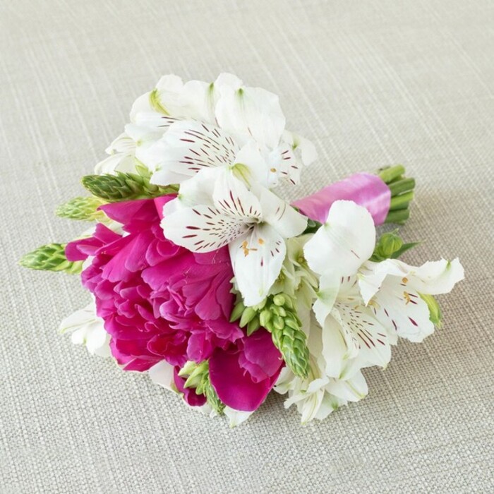 A richly coloured, compact floral piece lies across a softly woven, light beige fabric, evoking the look of natural linen draped over a table in an Oxford townhouse or a small venue tucked off Cowley Road. At the centre of this design is a magnificent fuchsia peony, its petals tightly nested in multiple ruffled layers that build a plush, domed shape, like a silk cushion crafted from living colour. Around this central bloom, several white alstroemeria flowers open in different directions, their six slightly flared petals forming elegant trumpets. On the inner surfaces of these petals, fine maroon and deep red streaks and speckles radiate from the throat, introducing a delicate pattern that draws the eye closer, the markings appearing almost like quick brushstrokes from a careful hand. Slender, elongated green buds and spiky florets thread between the main flowers, injecting energy and movement into the arrangement while adding a bright, youthful green that contrasts beautifully with the bold magenta and crisp white. The stems are gathered in a neat, compact bundle and wrapped in a glossy satin ribbon in a vivid, matching pink, its smooth sheen catching the light and lending a polished, formal finish that suggests the work of an experienced local florist. The lighting is soft and even, creating gentle shadows under each petal and emphasising the textures of the blooms and the faint weave of the background fabric. The overall impression is of a small but luxurious boutonnière, corsage, or intimate bouquet, ideal for weddings and special occasions across Oxford-from elegant ceremonies near Christ Church Meadow to relaxed celebrations in Jericho-capturing a blend of modern vibrancy and classic romance.