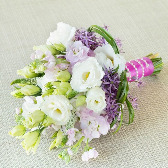 A strikingly elegant, hand-tied bouquet rests diagonally on a tactile, softly textured linen or burlap fabric, its neutral beige echoes the understated refinement of Oxford's timeless architecture. Several prominent white lisianthus, with luxuriously ruffled petals and light yellow-green centers, form the bouquet's luminous core-symbols of purity and new beginnings. Alongside these, clusters of small lavender-pink flowers, likely stock, intermingle to add softness and gentle perfume, creating a dance of pastel hues. The deep purple of ornamental allium introduces dramatic contrast, their star-shaped florets forming spiky bursts of color that enliven the design and suggest the dynamism of Magdalen College gardens in late spring. Nestled throughout, bright lime-green buds and elegantly textured green fillers provide a sense of vitality and dimension, their feathery presence reminiscent of wild botanicals found near Port Meadow. Slender arching bear grass blades frame the ruched flowers with a sense of movement, binding the composition together. A magenta satin ribbon tightly embraces the stems, its exuberant color highlighted by the ethereal touch of a white pearl strand-lending a sense of occasion and classic Oxford celebration. This bouquet's harmonious palette of white, lavender, purple, and fresh green makes it a perfect choice for weddings, anniversaries, or sophisticated gatherings near the heart of Oxford. It reflects a local tradition of grace and artistry, with freshness and elegance tailored for delivery by Flowers Oxford.