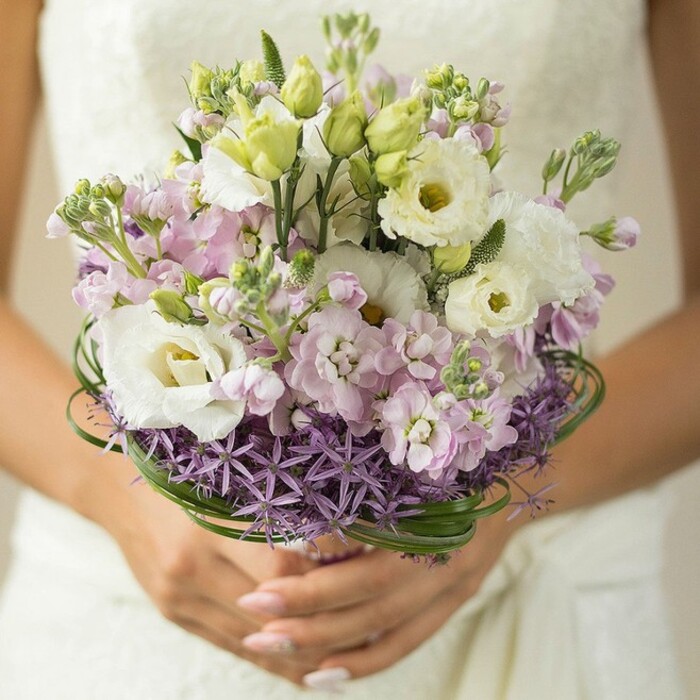 This close-up wedding bouquet from Oxford radiates elegance and invitation, captured at the moment a bride gently cups the sturdy stems, her nails polished in a soft nude or barely-there pink. Behind, the faint, lace-edged outline of a white or ivory gown blurs into the atmospheric Oxford light, ensuring that the flowers themselves remain the focus. At its heart, the bouquet features lush white Lisianthus with their signature ruffled petals and subtle centers tinged with green and yellow-flowers often cherished by local Oxford florists for their understated grace. Interspersed among these are airy tufts of light pink and lilac stock, their petals loosely layered to create a soft transition and tactile sense of depth, reminiscent of a spring walk through University Parks. Fresh, unopened Lisianthus buds highlight the arrangement's natural vigor, while a solid ring of spiky, star-like purple Allium forms a modern, architectural base, a nod to Oxford's diverse botanical traditions. Elegant, slender bear grass is looped above the Allium, adding a sculptural frame of deep green that both grounds and elevates the pastel blooms. The result is a vibrant yet gently harmonious composition, perfect for ceremonies set in Oxfordshire's historic halls or riverside gardens-a bouquet that blends contemporary floral artistry with classic imagery, celebrating romance and local craftsmanship.