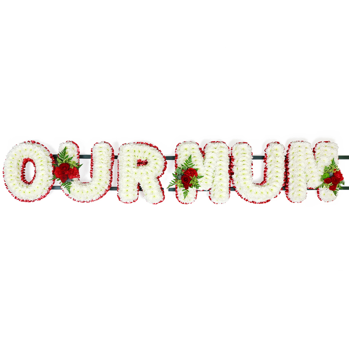 Set against a spotless white background, this OUR MUM funeral lettering tribute appears calm and composed, as though ready to be placed in a peaceful corner of an Oxford church or laid beside a resting place in one of the city's historic cemeteries. The words are formed from large block letters, each letter crafted with meticulous precision and densely filled with tiny white chrysanthemum heads or similar soft blooms. Their overlapping petals create a velvety, cushion-like surface that looks both gentle and substantial, suggesting the closeness and care of family. Around the outer edge of every letter runs a fine, continuous border of deep red blossoms, likely spray carnations or compact red flowers, offering a powerful contrast that sharpens the outline and visually holds the design together. Three small but striking rose sprays punctuate the display: one spray nestles between the O and U, the second lies between the U and the first M, and the third graces the far right, touching the final M. Each cluster features fully opened red roses surrounded by lush green leaves and wisps of fern, the colours evoking deep love, respect, and remembrance. A dark green horizontal bar subtly links the letters from behind, hinting at a sturdy frame that makes the tribute suitable for transport and display during a funeral procession through areas like Headington, Summertown, or Cowley. The overall impression is one of quiet elegance and heartfelt sentiment, a bespoke floral message that would feel at home in any Oxford memorial setting, expressing devotion, gratitude, and a tender goodbye to a much-loved mum.