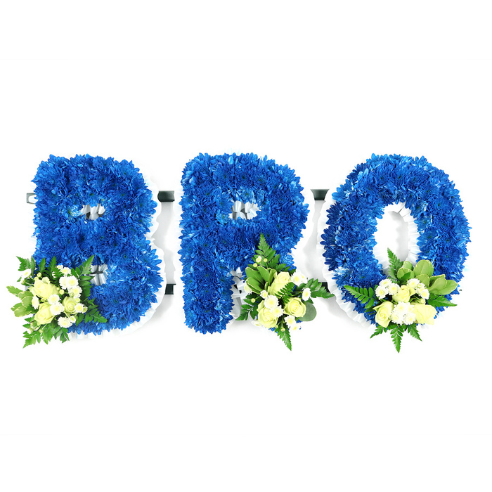 An intimate studio portrait of a three-letter sympathy tribute reading BRO, crafted in dense royal blue chrysanthemums against a pure white background. Each letter is freestanding, built on a slim white foam frame that peeks through at the inner loops and edges, revealing the careful structural work behind the floral surface. The B, R and O are entirely covered with saturated blue chrysanthemum florets, their massed petals creating a sumptuous, tactile quality that is at once dignified and consoling. Subtle floral accents sit at the base of each letter: clusters of cream or pale yellow spray roses, some small and tight, others softly opening, interspersed with delicate white filler blooms and a mix of greenery from glossy, wide leaves to airy fern-like stems. The controlled studio lighting highlights the chromatic contrast between the deep blue and the light, creamy accents, while casting soft shadows that lend dimensionality without detracting from the arrangement's solemn purpose. This composition reads as a quiet, handcrafted declaration of love and memory-suitable for placement beside a photograph or at a graveside service-and would be appropriate for families arranging tributes in Oxford, whether near Magdalen Bridge, the High Street chapels, or at a city cemetery. The overall effect is respectful, personalised and thoughtfully made to honour a brother's life.