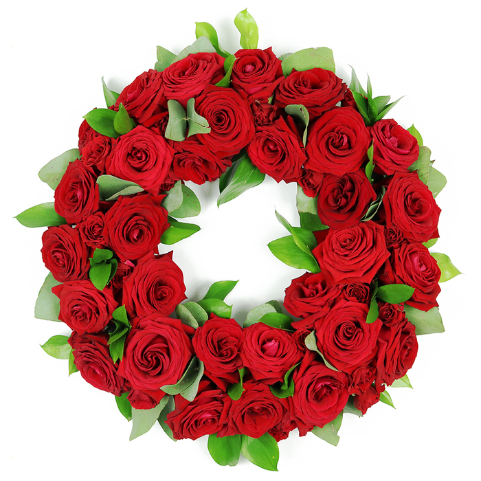 A deeply symbolic and visually striking circular wreath, meticulously crafted from dozens of vibrant, deep red roses, rests against a pristine white background. The wreath is densely packed, displaying a lush, opulent tapestry of crimson blooms at varying stages of opening, from tightly furled buds to elegantly unfurled, velvety petals, each displaying rich hues and intricate folds. These abundant red roses, traditionally symbolizing love, remembrance, and respect, form a continuous ring. Interspersed are clusters of fresh, glossy green foliage. Some leaves are broad and rounded, possibly eucalyptus, while others are slender and pointed, offering verdant contrast that deepens the arrangement. The careful layering gives the wreath a three-dimensional quality, with petals and leaves overlapping gracefully. This elegant red rose wreath conveys solemn beauty, heartfelt emotion, and enduring tribute. It is a powerful visual element, often associated with memorial services, funeral arrangements, expressing sympathy and condolences, or honoring anniversaries. The striking combination of passionate red and serene green against a white backdrop makes this wreath a poignant symbol of remembrance and respect in Oxford's communities, from Port Meadow to Oxford city centre, and beyond. It sits as a quiet testament to memory, an offering of condolence that is both timeless and contemporary. When placed in a church, chapel, or at a family home, it speaks with dignity across a room full of mourners.