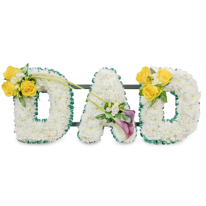 A striking, handcrafted memorial display spelling DAD, each letter constructed from a dense bed of immaculate white spray chrysanthemums that create a smooth, rounded block form with rich textural depth. The edges of every letter are crisply outlined in a ruffled dark green satin ribbon, producing a deliberate line that frames the white blooms and adds a subtle sheen beneath soft lighting. Behind the letters thin, dark green metal bars run horizontally, visible in places and serving as the sturdy spine that binds the three letters into a single ceremonial panel. The left D is accented on its upper left curve with a concentrated cluster of bright yellow roses-about three to four open heads-combined with tiny white freesia or gypsophila and mixed greenery; two slender, bare green stems arch diagonally toward the centre, introducing movement. On the A a composed grouping sits on the lower diagonal: two deep purple-burgundy calla lilies provide an elegant, architectural counterpoint to the soft chrysanthemums, surrounded by small white accent blooms and glossy foliage. The right D mirrors the left, echoing the yellow rose cluster and leafy tendrils to create balance. Shot against a neutral white background with soft, directional light that emphasizes texture and shadow, the piece suggests a faint clean floral perfume and meticulous craftsmanship. Suitable as a sympathy tribute for services across Oxford-whether delivered to a chapel near Broad Street or a wake in Jericho-this DAD arrangement reads as respectful, expertly made, and quietly consoling.