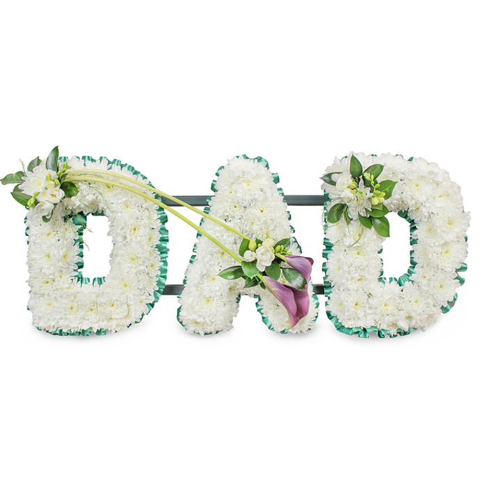 A poignant floral arrangement forms the word DAD in bold, block-style letters, each one three-dimensional and standing proudly against a clear white background that highlights the craftsmanship. The letters are completely packed with countless white chrysanthemums, creating a smooth, plush surface of tiny, overlapping petals that look almost like fresh snow. This uniform white canvas conveys purity, respect, and a gentle stillness, much like the reflective atmosphere at a funeral service in one of Oxford's historic churches. Around the outer edge of every letter runs a continuous ribbon of deep emerald green satin, carefully pleated to produce a ruffled border that defines and frames the shapes. The ribbon's sheen catches the light softly, echoing the natural green found in Oxford's college gardens and along the towpath by the river. Subtle glimpses of a dark metal frame and horizontal bars appear behind the letters, confirming that this is a standing tribute, designed to be placed beside the coffin or at the front of a chapel. The first D is further softened by a small posy placed on its upper left curve: delicate white freesias with slightly open trumpets and closed buds, nestled among fresh green foliage. Two long, arching green stems extend from this area, sweeping diagonally across to meet the lower right side of the central A, where they form an elegant focal spray. Here, two deep purple calla lilies, with their sculpted, trumpet-shaped blooms, rest among more white flowers and rich green leaves, bringing a dignified touch of colour and depth. The final D on the right carries a smaller, matching cluster of white freesias and greenery on its upper right edge. The composition feels deeply personal and tender, a classic tribute a family in Oxford might choose to honour a much-loved father with grace and quiet emotion.