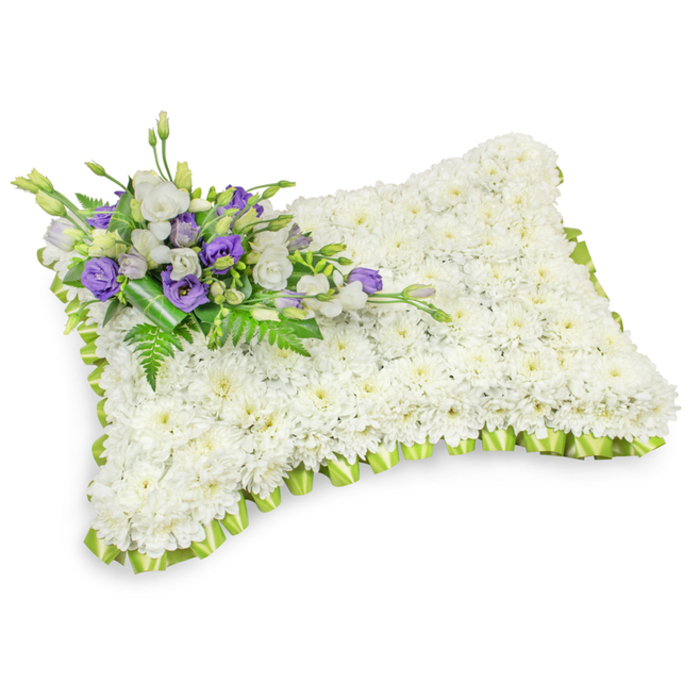 An exquisitely shaped sympathy pillow fashioned from a dense carpet of small white chrysanthemums, each head arranged to form a plush, unbroken field of ivory that reads like a soft blanket of flowers. The rectangular form is gently rounded at the corners and slightly domed, so the tribute sits with a comforting, pillow-like presence; under studio light the petals catch soft highlights and the shallow shadows beneath the piece give it a tangible, three-dimensional weight. The entire perimeter is finished with a pleated light-green satin trim, a subtle, fresh border that delineates the tribute's shape and introduces a whisper of springtime renewal. Across the top-left a composed bouquet of accents brings thoughtful contrast: pristine white lisianthus paired with deeper purple lisianthus - some opening fully, some held in slender buds - together speak of respect and remembrance. Sprays of feathery fern fronds and broad dark leaves are woven through the cluster, and a neat wrap of deeper green foliage secures the stems, lending the arrangement a refined, handcrafted look. The fragrance is soft and green, the chrysanthemum providing a clean, almost tea-like scent while the lisianthus contributes a gentle, floral note. I envision this piece arriving at a funeral parlour in Botley Road or being placed at a family service near University Parks, where understated, elegant tributes are preferred. Prepared with care for local services across Oxford, this pillow offers a composed, tender way to express sympathy when words are not enough.