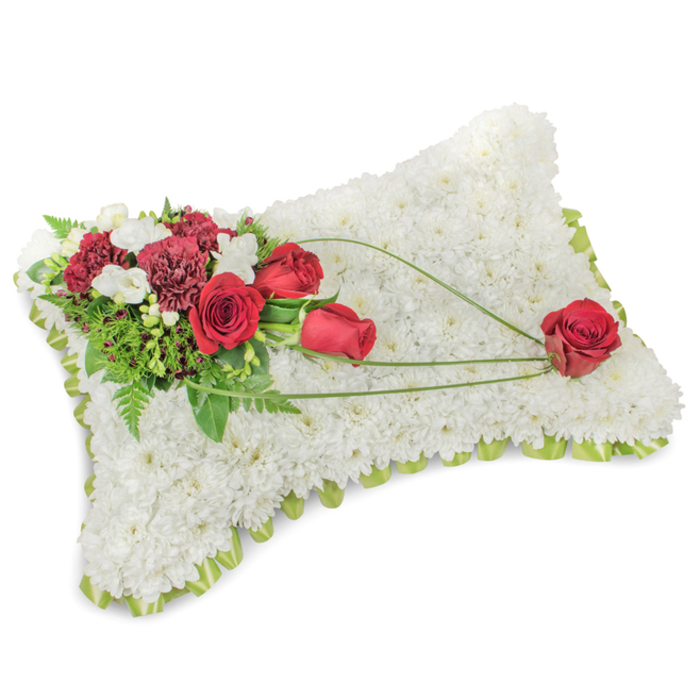 An exquisite rectangular pillow-style tribute, the face entirely covered with a dense carpet of small, immaculate white chrysanthemums that together create a soft, downy texture resembling a restful blanket of petals. The slightly raised edge reveals a hint of pale green ribbon or delicate foliage that frames the piece with a whisper of colour, lending a finished, hand-crafted air. On the upper left an abundant spray of deep red roses and rich burgundy carnations forms the principal visual anchor; the roses present both closed buds and fully unfurled blooms whose velvety surfaces catch the light, while the carnations' frilled petals add sculptural interest. Intermingled are little clusters of white freesias or similar tiny blooms that bring brightness and gentle perfume, and a lush assortment of greenery-fine fern fronds and denser leaves-grounds the composition. From that corner a set of slender green stems or bear-grass arcs sweep diagonally across the white chrysanthemum field, trailing toward the lower right and finishing at a single, immaculate red rose that appears cradled by the curving foliage. The arrangement's tones-pure white, bold crimson and fresh green-work together to suggest purity, enduring love and quiet respect. In the soft morning light of an Oxford chapel or along the High Street near Radcliffe Camera this piece radiates calm and dignity. It is often selected for funeral services or memorial displays, carrying a measured floral scent and a handcrafted feel that many families in Cowley and Headington find comforting.