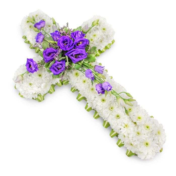 In this image, a traditional Christian cross-shaped funeral tribute is captured from a slightly elevated angle, resting on a pristine white background that enhances its sense of calm and solemnity. The entire form of the cross is meticulously packed with small white spray chrysanthemums, each blossom sitting closely against the next to create a plush, almost quilted surface. Their rounded petals form a gentle texture that calls to mind the quiet stillness of early morning mists over Port Meadow in Oxford. You can see the faint green centres of the chrysanthemums, adding a delicate sprinkle of colour within the sea of white. Along the edges of the cross, a tidy band of vivid lime-green ribbon traces the outline, offering definition and a fresh botanical accent, as if outlining the silhouette with new spring growth. At the heart of the cross, a rich focus of deep purple lisianthus blooms commands attention. The lisianthus are lush and fully open, with layered, ruffled petals that create depth and shadow, symbolising reverence and respect. Intertwined among these central flowers are slender shoots of soft green foliage and a few unopened buds, lending a natural, garden-like feel. Some of the lisianthus stems extend gracefully from the main cluster, drifting down the lower right arm of the cross in a gentle diagonal, adding a sense of movement and flow. The lighting is soft and even, casting only a modest shadow beneath the tribute, ensuring the colours remain true and the intricate workmanship is clearly visible. Often chosen for services in local churches from Cowley to Summertown, this floral cross embodies sympathy, Christian faith, and enduring remembrance.