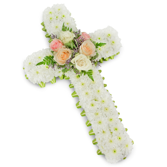 A reverent white floral cross dominates the image, presented against a bright studio-style white backdrop that lets the intricate floristry stand out with clarity. The cross itself is built up with an abundance of small white chrysanthemum button heads, each bloom pressed closely into the next so the entire front surface appears padded and soft, almost like a quilt of petals. This dense, uniform covering conveys a feeling of purity, peace, and gentle stillness, the kind of calm one might find walking through an old Oxford churchyard on a quiet afternoon. Running neatly around the outer contour of the cross is a narrow band of lime green ribbon, visible just beyond the petal edge. This subtle border sharpens the silhouette and adds a crisp freshness, reminiscent of new leaves in University Parks as spring begins. At the central meeting point of the arms and the vertical stem, a carefully arranged cluster of roses draws the eye. These roses, fully open and velvety, display harmonious shades of ivory, peach, and soft blush pink. They are interwoven with delicate fern fronds that arc lightly outward, and scattered sprigs of pale lavender fillers that bring a hint of colour contrast without breaking the overall softness. The composition feels balanced and intentional, each element supporting the symbolism of faith, remembrance, and enduring affection. This floral cross would be entirely at home at a funeral or memorial service in Oxford, perhaps placed near the lectern of a city-centre church or beside a photograph of the departed at a crematorium, quietly expressing sympathy and spiritual hope.