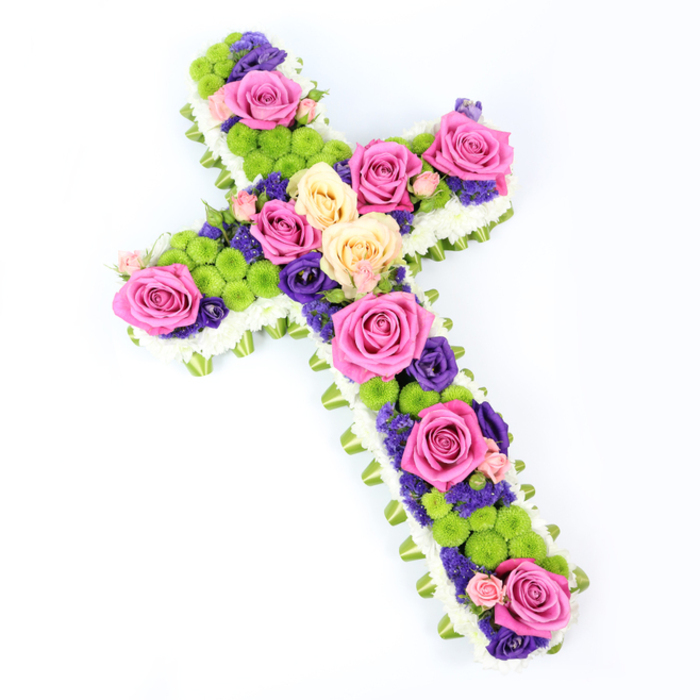 A beautifully composed floral cross rests against a luminous white background, shown from a slightly overhead angle so that its structure and intricate detailing can be fully appreciated. The traditional shape of the cross is crisply outlined with a thick edging of white spray chrysanthemums, each tiny bloom nestled tightly next to its neighbour, creating a continuous, cloud-soft frame. Peeping out just beyond this white border is a slender band of light green ribbon or folded foliage, forming a subtle yet sophisticated trim that lends depth and a gentle hint of garden freshness, not unlike the greenery lining Oxford's historic college quadrangles. Inside this delicate frame, the cross is filled to overflowing with richly coloured flowers. Large roses, their petals fully unfurled, appear in radiant hot pink and softer shades of creamy pale yellow, positioned thoughtfully along the arms and stem to guide the gaze and create natural focal points. Around and between these roses sit rounded clusters of lime-green button chrysanthemums, their almost spherical heads adding lively texture and a modern flair. Woven throughout are smaller purple and violet flowers, probably statice or lisianthus, whose deeper tones bring a sense of calm, reverent depth, similar to the quiet corners of churches around the city centre. The entire design is angled, tilting gently from the upper left to the lower right, which gives the impression of movement and dimensionality, as if the flowers are lifting slightly from the background. Bathed in clear, even light, every colour appears true and vibrant, and every petal edge is visible. This floral cross evokes faith, remembrance and comfort, making it a fitting funeral or memorial tribute for families gathering in Oxford to honour a loved one.