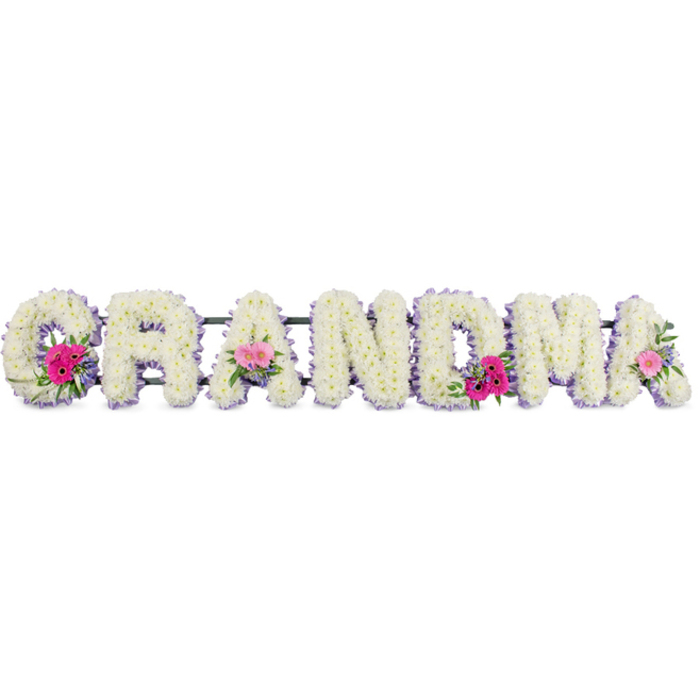 In this photograph, the word GRANDMA is written not with ink, but with flowers, each letter formed from an abundance of white chrysanthemums that create a plush, cushion-like surface. The blossoms are tightly clustered, their short stems hidden from view so that only layers of velvety petals remain, suggesting softness and purity. Around each letter runs a slender band of lilac ribbon, its muted purple tone adding a delicate contrast and discreetly defining the outlines. The satin finish catches the light, giving a subtle shimmer that brings a hint of elegance without overpowering the serenity of the design. Set along the sequence of letters, three small, rounded posies add brighter notes of colour: on the first G, one of the central As, and the final A. These accents feature cheerful pink gerbera daisies, their large central discs surrounded by smooth petals, as well as soft pink roses and short lengths of glossy green foliage. The foliage fans out and curls naturally, evoking the gentle abundance of an Oxford garden in spring or a quiet corner of Christ Church Meadow. The arrangement sits under even studio lighting, which highlights the fresh texture of the chrysanthemums, the subtle blush of the roses, and the clean line of ribbon, while leaving a calm, uncluttered background. Though the image is still, it suggests movement through memory-of stories shared, walks through Oxford's historic streets, and moments at family tables. The tribute feels personal and respectful, a piece likely created by a local florist who understands how to balance comfort, tenderness, and formality in a farewell for a cherished grandmother.