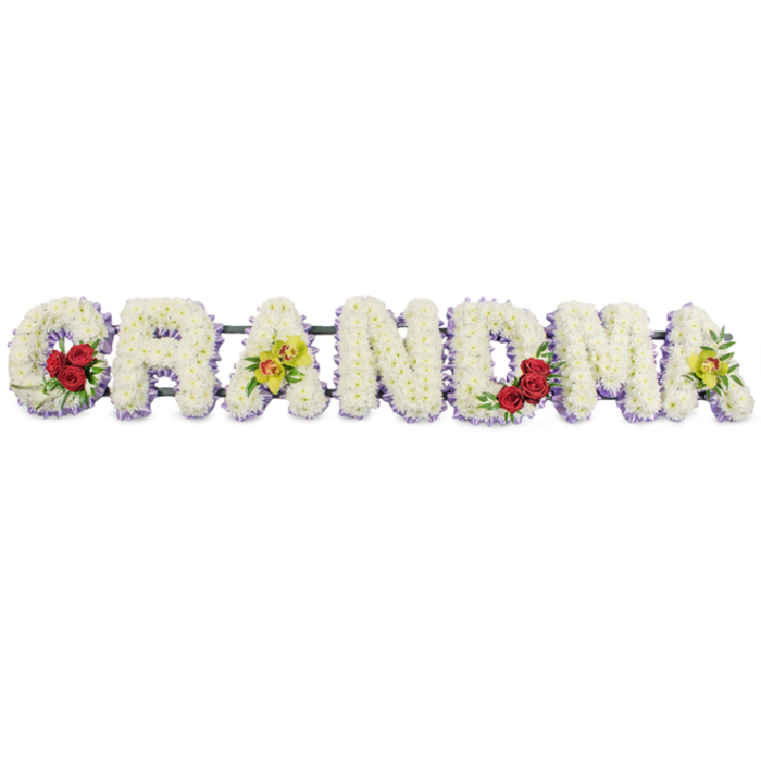 A striking floral word tribute spelling GRANDMA dominates the image, the letters set in a single horizontal line against an uncluttered white backdrop so the flowers take centre stage. Each capital letter is carefully shaped and then completely covered with small white chrysanthemums, or similar button flowers, packed so closely that no gaps of foliage or frame show through. Their tiny petals create a velvety, uniform texture, giving the impression of pure snow moulded into letter forms. Underneath this white blanket, a trim of pale lavender or light purple blooms peeks out around the base of each letter, forming a soft outline that lifts the word away from the background. Carefully placed accent flowers personalise the design. On the lower left curve of the G, three luxurious red roses are gathered together among glossy, deep green leaves, drawing the eye to the start of the tribute. The first A from the left displays two vivid yellow-green orchids with speckled reddish throats laid across the inner bar, their slightly waxy petals adding contrast in both colour and texture. The R, N, and D are kept simple and serene, composed solely of the white blooms and lavender edge, allowing the coloured accents to remain special. The M features another small group of red roses nestled into its lower right arch, while the final A echoes the first with two more yellow-green orchids resting in its open centre. A discreet dark metal frame unites all the letters along the back, ready to sit on an easel or coffin spray stand. It is the kind of carefully considered memorial arrangement a local Oxford family might have delivered to a funeral at Wolvercote or Botley, offering a tender, visually striking farewell to a beloved grandmother.