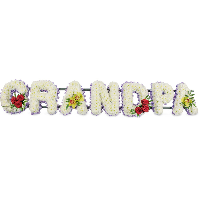 A poignant funeral arrangement spells out the word GRANDPA in large, block-style capital letters, stretched horizontally against a stark white backdrop that brings every bloom into focus. Each letter is densely massed with pristine white chrysanthemums, their countless small petals overlapping to create a plush, cushion-like surface that feels soft, gentle, and reverent, as if inviting a final, loving touch. A slim band of pale purple or lavender ribbon traces the perimeter of each letter, adding a delicate outline that stands out just enough to define the word without disturbing its calm simplicity. In the sweeping curve of the opening G, a compact cluster of deep red roses rests among green foliage, the velvety petals providing a dramatic contrast against the white, like a vivid memory held close in the heart. The third letter, A, introduces a refined spray of yellow-green orchids, with two or three intricate blooms lending an air of quiet elegance and individuality, reminiscent of a cherished grandfather's unique character. The rounded bowl of the central D echoes the G with another grouping of red roses, maintaining visual rhythm, while the final A on the right side is again graced with a small spray of orchids, bringing the tribute into gentle visual harmony. Slim, unobtrusive horizontal bars run behind the letters, keeping them aligned and secure, ready to rest beside a coffin or at a memorial gathering in Oxford, perhaps in a church near Jericho or a chapel out in Kidlington. This thoughtful combination of pure white flowers, romantic red roses, and graceful orchids forms a tender, personalised farewell, honouring a beloved grandfather with both dignity and heartfelt emotion.
