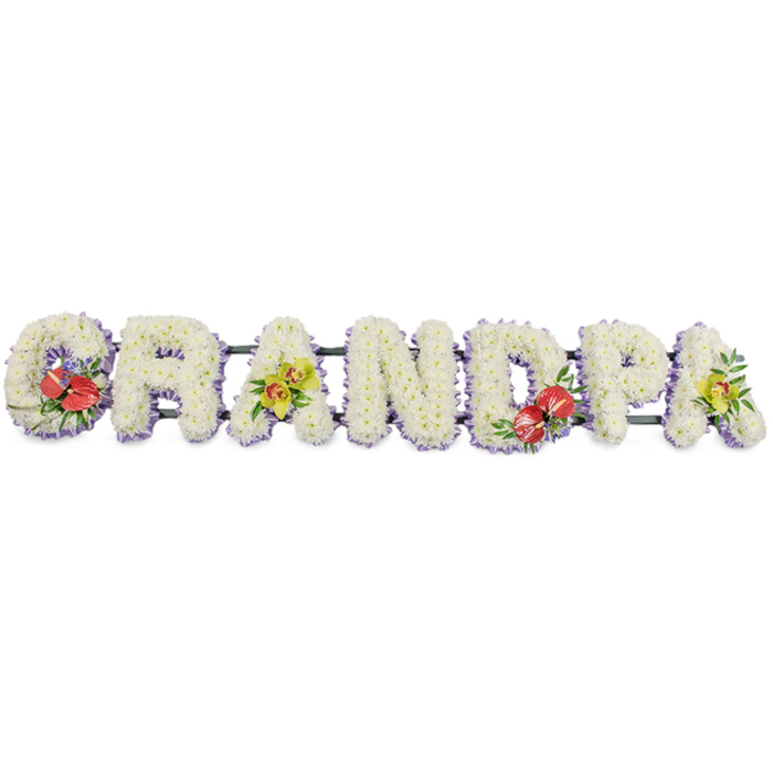 This photograph captures an elegant funeral flower arrangement spelling the word GRANDPA in bold block capitals, designed as a long horizontal tribute that could rest along the base of a coffin or at the front of a chapel in Oxford. Each letter is thick and solidly built, completely covered with masses of small white chrysanthemums whose petals overlap to create a plush, velvety surface. The blooms are pristine, suggesting they have been freshly prepared by a florist on the morning of the service, with each individual flower carefully angled to hide the mechanics beneath. Around the lower edge of each letter, a narrow band of pale lavender ribbon peeks out, forming a delicate border that outlines the shapes and introduces a soft hint of colour without disturbing the overall white serenity. A dark green bar runs discreetly behind the letters, supporting the tribute so it can be moved easily between the hearse, the service in an Oxford church, and the final resting place. Four letters are accentuated with richer tones. The 'G' and 'D' carry bold clusters of deep red anthuriums, their distinctive heart-shaped, glossy spathes and central spadices standing proudly against a backdrop of dark green leaves and a few purple-toned fillers. On the first and last 'A', sprays of lime-yellow orchids cascade lightly over the chrysanthemums, their refined, arching stems and open blooms bringing lightness and grace. Together these colours convey affection, respect, and remembrance for a much-loved grandfather, making the tribute a fitting floral farewell for families gathering anywhere from central Oxford churches to more secluded village cemeteries nearby.