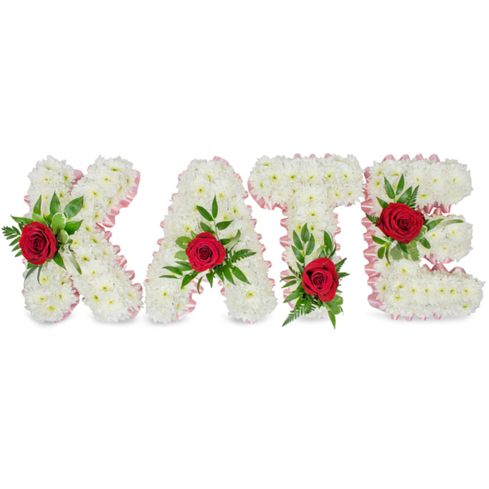 A striking, personalized floral tribute spells KATE in prominent three-dimensional block letters, each letter densely covered in crisp white chrysanthemums that form a voluminous, cloud-like surface. A delicate pale pink satin ribbon trims the contours of every letter, its soft sheen lending refinement and a gentle blush to the overall palette. Each letter is accented with a single, richly coloured red rose set amid a generous spray of green foliage - a combination of glossy leaves and delicate fern fronds that give the piece natural texture and depth. The florist has positioned the roses with intention: on the K the rose nestles low along the left vertical stroke, the A bears its rose at the very peak of the triangular top, the T is emphasised by a bloom at the base of the left side of its crossbar, and the E carries its rose on the middle horizontal arm. Presented against a pristine white background, the arrangement reads as a composed and dignified memorial, the white chrysanthemums conveying purity, the red roses declaring love and admiration, and the green suggesting resilience and renewal. Created with the sensibility of a local studio, this bespoke tribute is the kind of piece families in Oxford might request for services at Christ Church Meadow, a small chapel, or a celebration of life. Flowers Oxford crafts such lettered tributes with attention to scent, texture and proportion, and can arrange delivery across Headington, Jericho and Summertown to suit funeral or remembrance plans.