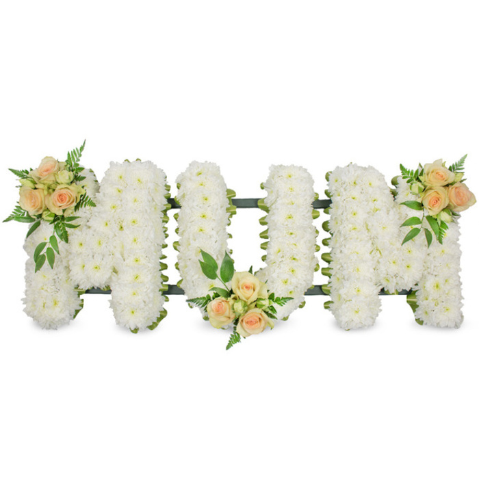 This photograph shows a long, horizontal funeral flower tribute spelling out MUM in large, upright block capitals, presented crisply against a bright white background to focus attention on the detail of the floristry. Each of the three letters is meticulously filled with compact white spray chrysanthemums, their small daisy-like flower heads pressed close together to create a smooth, plush surface without any visible gaps. The uniform white blooms form a serene backdrop, their soft texture suggesting gentleness and tender care. Slight glimpses of dark green support work or ribbon trace the edges of the letters, subtly outlining the shapes and providing a fine, neat frame that keeps the design clean and structured. Resting on this pure white foundation, three soft, romantic clusters of roses bring colour and personality. At the top left of the first M, a carefully arranged group of peach and blush pink roses, some in bud and some half-open, is cradled by sprays of lush green foliage and delicate ferns. A second cluster of the same warm-toned roses decorates the lower centre of the U, drawing the eye naturally along the arrangement. The final rose accent is placed on the top right of the second M, giving the tribute a balanced, symmetrical feel from one end to the other. The overall impression is of a bespoke, heartfelt memorial piece, the kind of personalised MUM tribute that might be chosen for a service at Oxford Crematorium or a quiet farewell in a village church near Cowley or Iffley. The combination of crisp white chrysanthemums and soft peach roses conveys respect, deep affection, and enduring remembrance for a much-loved mother.
