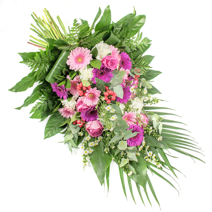 Against a bright white backdrop, a striking asymmetrical funeral spray is arranged in a graceful diagonal, starting full and abundant at the upper left and gradually tapering to a cascading trail of foliage at the lower right. The eye is immediately drawn to the cluster of gerbera daisies in the centre, their large, round faces blooming in shades of blush pink, bold fuchsia, and rich magenta. These are paired with refined pink roses, their gently spiralled petals adding depth and a sense of quiet romance, much like the soft floral displays often seen in Oxford's historic college chapels. Around them, clusters of ruffled white flowers-likely mini carnations or dianthus-create a cloud-like contrast, brightening the arrangement and adding comforting texture. Tiny white daisy-style filler flowers are threaded throughout, giving a light, airy feel that suggests hope amidst sorrow. Nestled among the petals are vivid red hypericum berries, their smooth, bead-like forms providing little sparks of colour and a natural, woodland charm. The spray is anchored by a generous base of mixed greenery: broad, glossy dark leaves form a supportive backdrop, fine fern fronds introduce movement and softness, and silvery eucalyptus branches lend both visual freshness and a hint of cool fragrance. From the lower right corner, long, sword-shaped palm leaves extend outward, enhancing the sense of flow and direction. At the top, the gathered green stems are clearly visible, showing the skill of the florist who has bound them. This dignified tribute could rest on a coffin or an easel at a funeral in or around Oxford, offering a heartfelt expression of sympathy, remembrance, and peaceful farewell.
