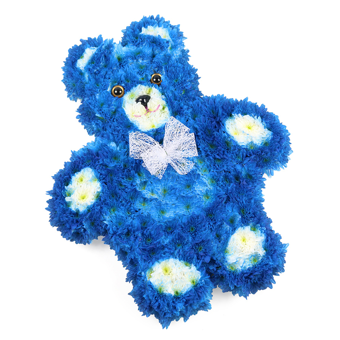 A three-dimensional teddy bear made entirely of fresh flowers rests on a clean white background, its form so carefully shaped that every curve reads clearly, even without colour. The bear's main body is a deep royal blue, composed of countless small, tight-petalled blooms that sit shoulder to shoulder, mimicking the dense nap of a favourite plush toy. Each petal catches soft, diffused light, creating subtle shadows that suggest depth and softness rather than rigid structure. In gentle contrast, clusters of creamy white and pale yellow flowers define the inner cups of the ears, the rounded muzzle, and the four oval pads on the paws, as though the teddy is wearing tiny, light-coloured mittens and booties. The eyes are smooth and round, a warm brown with darker pupils that seem to gaze kindly outward, while the little black triangle nose and a neat curved mouth line give the bear a peaceful, almost protective expression. Around the neck, a fine white mesh bow is tied with care, its translucent loops and tails resting lightly across the chest, adding a touch of grace and ceremony. The composition is photographed from above with a gentle diagonal tilt, making the bear appear as if it has just been laid down with love. As a florist working in Oxford, I see this style of floral teddy chosen by families from Kidlington to Iffley who wish to honour a child or baby, or to reflect the soft-hearted nature of someone remembered. The cool blue tones can evoke the tranquillity of the Thames by Port Meadow at dusk, while the overall design offers comfort, remembrance, and a quiet embrace expressed through flowers.