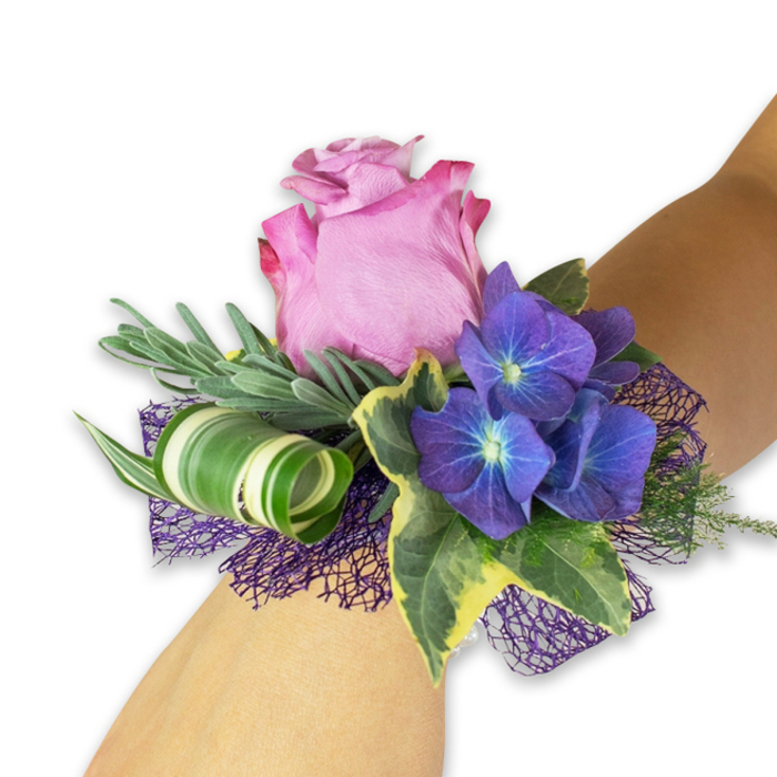 Shown on a pale forearm against a clean white field, this wrist corsage blends classic romance with contemporary detail in a way I often make for brides and prom groups arriving from Jericho and Summertown. Its centrepiece is a large, partially open pink rose with layers of silky petals-lighter toward the middle and graduating to a deeper magenta around the ruffled margins-creating a soft, inviting focal point. Surrounding the rose are three prominent clusters of deep indigo-purple hydrangeas whose minute florets carry subtle blue-white centres, giving the clusters a luminous, almost starlit texture that balances the rose's warmth. The design is anchored by silvery-green, narrow sprigs (reminiscent of eucalyptus or lavender) that add a muted, chalky green softness and a faintly aromatic note, while glossy variegated ivy leaves with dark green hearts and creamy-yellow edges introduce a traditional, garden-like elegance. A bold, wide leaf striped in white and green has been artfully curled into a tight spiral to provide a modern, sculptural counterpoint, and beneath this natural architecture a dark purple open-weave mesh peeks through, enhancing depth and echoing the hydrangea tones. As a local florist at Flowers Oxford, I choose these elements to combine tactile contrasts-the silky rose, papery hydrangea, crisp ivy and soft mesh-so the wearer feels both celebrated and comfortable whether walking through the Covered Market or posing for photographs in the Botanic Garden.