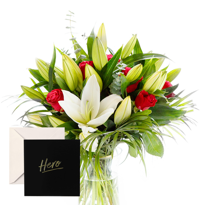 As a florist, there is joy in blending the bold with the serene, and this arrangement embodies that harmony. The white lilies drift at the heart, luminous and airy, their satin petals contrasting magnificently with the tactile, lush greenery that encircles them. Rich ruby roses punctuate the design, each one a symbol of love and respect, their fragrance mingling softly with the lilies in the overall aromatic chorus. The verdant leaves provide a cooling, forest-like backdrop, while unopened lily buds hint at anticipation and new beginnings and the subtle promise of growth. In soft, natural light the bouquet feels alive and uplifting, a fitting tribute for honoring someone's strength or celebrating a significant achievement. The arrangement carries a sense of quiet grandeur, with careful balance and generous spacing that allows the eye to travel from lily to rose to leaf. A delicate trail of wispy stems adds movement, while the scent of green and bloom lingers pleasantly in the room. The accompanying card marked Hero makes the gesture feel intimate, enduring, and deeply personal. Born from a trusted Oxford florist, this bouquet is built to hold its own in a busy hallway or at a ceremony table. It travels well, arriving fresh and vibrant for citywide delivery across Jericho, Osney, Headington, and beyond, offering a graceful way to recognize resilience, achievement, or heartfelt gratitude in the heart of Oxford.