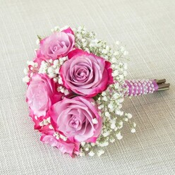 Nestled atop a softly textured, taupe-hued linen, this graceful bouquet evokes the timeless romance of an Oxford wedding. At the heart of the arrangement are radiant pink and lavender roses, each petal displaying a captivating gradient that moves from gentle purple interiors to rich magenta and fuchsia edging. Intertwined throughout are frothy clusters of white baby's breath-known locally as gypsophila-which introduce an ethereal and airy contrast to the velvety fullness of the roses. The bouquet's stems are neatly aligned, encased in a sheen of deep purple satin ribbon, and finished with iridescent white pearls spiraling in a perfectly parallel formation-bringing to mind the refined atmosphere and gentle pageantry of Oxford's many traditions. From the lush green stems that peek from beneath the wrapping to the fresh, uplifting scent that hints at spring across Christ Church Meadows, every detail whispers of celebration and attention to detail. Crafted by an experienced local florist, this bouquet is perfectly suited for brides, bridesmaids, or as a centrepiece for special anniversaries and events in Oxford, infusing every occasion with a sense of heritage and contemporary elegance.