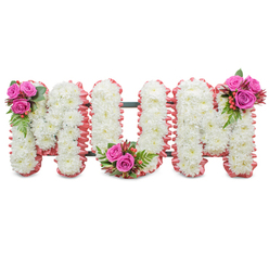 A reverent floral tribute spelling out "MUM" in large, connected capital letters, lovingly constructed from dense clusters of immaculate white chrysanthemums. Up close, the small chrysanthemum heads form a near-continuous, cloud-like surface across each letter, their petals creating a delicate, textured field that speaks of purity and calm. Each letter's perimeter is softly edged with a ruffled blush-pink ribbon or fabric trim, introducing a comforting warmth against the white blooms, while the dark green structural frame between the letters remains subtly visible to support the standing tribute. Three coordinated accent bouquets interrupt the white with bright, emotional colour: a compact spray of deep fuchsia-pink roses, fern-like greenery and glossy reddish-orange hypericum berries sits at the upper left of the first 'M'; the same combination nestles into the base curve of the 'U'; and a final mirrored cluster decorates the upper right of the second 'M'. The roses' velvety petals, the crisp fern fronds, and the shiny berries give pleasing tactile variety beneath gentle indoor lighting you might find in a chapel near Summertown or a family home around Iffley. This is the kind of arrangement families from across Oxford - from Jericho to Headington - request when they want a personalised, heartfelt floral message for a funeral or memorial. Prepared with the care you'd expect from a local florist such as Flowers Oxford, the piece balances classic sympathy symbolism with subtle colour to honour a beloved mother with dignity and affection.