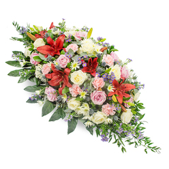 A refined double-ended funeral spray stretches diagonally across a clean white backdrop, its colours and textures echoing both sorrow and gratitude, like a thoughtful farewell arranged for a service in central Oxford. The eye is drawn first to the dramatic deep red lilies, their star-shaped blooms opening confidently, each petal gently recurved and dusted with darker markings. Around these striking centres, layers of roses in cream, soft white, and delicate blush-pink unfold, from firm buds to fully open rosettes, suggesting the many stages of a well-lived life. Light pink carnations, with their intricate, frilled petals, weave through the composition, while petite white daisy-like flowers and chrysanthemum sprays tuck into the spaces, creating a sense of abundance without heaviness. Cool-toned accents of blue-purple thistles and fine sprays of lavender-blue filler flowers introduce texture and depth, their slightly wild character balancing the more classical roses and lilies. The flowers are cradled in a bed of deep green foliage, a mix of glossy, broad leaves and softer, fern-like greenery that spills gently beyond the blossoms and tapers toward each end, ensuring the piece feels complete from all angles. The lighting in the image is soft and even, highlighting the natural sheen of the petals and the fresh vitality of the stems, as if the spray has just been prepared for same-day delivery to a family gathering near Headington or Summertown. The arrangement radiates quiet strength and compassion, a comforting tribute designed to honour cherished memories and offer solace at a moment of parting.