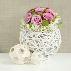 A meticulously crafted floral centerpiece rests on a pristine white tabletop, its design balancing contemporary style with soft romance, much like a quiet corner café off Oxford's Cornmarket Street. The arrangement forms a lush, domed bouquet emerging from a round, open-work vase made of interlaced white twigs, giving an airy, sculptural feeling. The main blooms are generously petalled roses in shades of soft pink and muted lavender, their outer edges touched by slightly deeper pink tones that hint at a just-unfolding bloom. Tucked between them, bright green button chrysanthemums create a lively rhythm of colour and texture, their compact, cushiony heads contrasting with the satin-smooth rose petals. Along the perimeter, tiny white and cream flowers weave in and out, like whispered accents, lending a gentle, cloud-like fringe that softens the structure. Beside the vase, on the same white surface, sit two decorative woven spheres in light, natural wood hues-one large, one small-mirroring the vase's crisscross pattern and reinforcing a theme of artisan craftsmanship. The background is a soft-focus expanse of beige fabric, subtly textured, reminiscent of linen napery laid for a wedding breakfast in an Oxfordshire barn. The lighting is diffused and flattering, revealing every curve of petal and twist of rattan without harsh shadow, ideal for showcasing wedding table flowers, chic home decor, or an elegant centrepiece designed for celebrations from Summertown to Headington. The entire image communicates care, freshness, and the thoughtful touch of a skilled local florist.