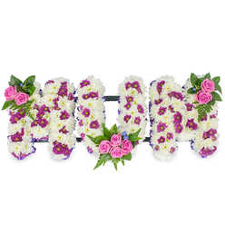 This sculpted floral tribute spells MUM in three substantial, three-dimensional letters, each formed from a plush base of immaculate white chrysanthemums that create a soft, cloud-like surface. Scattered through the white are clusters of deep magenta and purple spray chrysanthemums, their jewel tones adding shadow and visual rhythm across each letter. The arrangement is punctuated by vivid pink spray roses: a petite cluster sits at the upper left of the first M, a matching cluster adorns the upper right of the second M, and a larger, more prominent bunch of four to five roses is tucked into the lower curve of the central U, becoming the emotional centre of the piece. Lush dark green foliage - including feathery fern fronds and broader leaves - cradles the rose clusters, while tiny blue accent flowers slip in between blooms to cool the palette. A subtle, dark horizontal bar connects the letters, providing discreet structure so the tribute reads as a single, stable composition. The flowers catch gentle, diffused light, revealing the chrysanthemums' dense texture and the roses' velvety spirals; there is a soft, familiar scent of roses and green stems. As a local florist I picture this sympathy bouquet delivered with care to chapels near University Parks or to services around St Giles; at Flowers Oxford we craft each letter with the same respect and precise touch, making it suitable for a funeral, memorial or wake where family and friends gather to remember a mother with dignity.