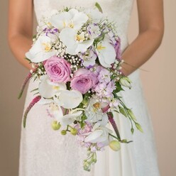 Bathed in gentle daylight, an Oxford bride poses with an opulent, cascading bouquet that captures the spirit of both celebration and serenity. Held delicately at her mid-torso, the bouquet flows in a waterfall style, highlighting proud white Phalaenopsis orchids whose broad, satiny petals catch every nuance of the natural light-much like the tranquil glow seen along Christ Church Meadow. Nestled around these orchids are lush roses in tender blush pink and soft lavender, their classic, velvety blooms hinting at cherished memories of spring weddings by the Botanic Garden. Wispy sprays of gypsophila breathe a graceful airiness through the arrangement, interspersed with petite, pale purple blossoms and snippets of verdant green leaf. Trails of vivid magenta amaranthus bring an unexpected texture, their tactile, draping forms resembling the historic willow trees beside the Cherwell. The bride's gown, glimpsed in soft ivory with delicate lace detail, completes the picture of timeless elegance. Above all, the pure white, romantic pink, and serene purple palette evoke a sense of calm anticipation, making the bouquet as perfect for Oxford's storied venues as for a sunlit garden ceremony. Glancing at this design, one is transported to a moment filled with hope, grace, and the quiet excitement that precedes a life of shared dreams amidst Oxford's enchanting landscape.